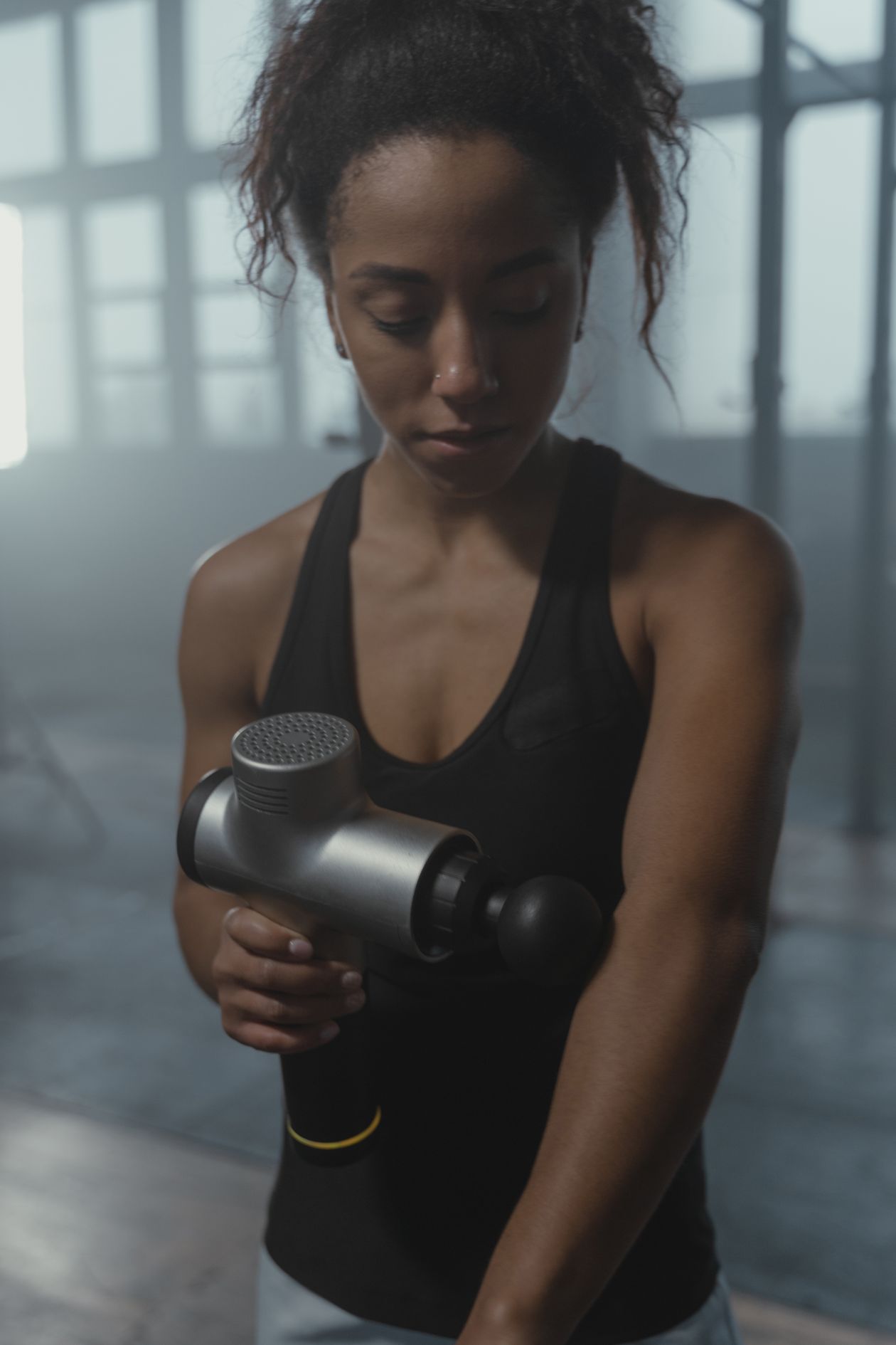 woman using a massage gun on her arm in a gym environment showing percussive therapy for muscle recovery