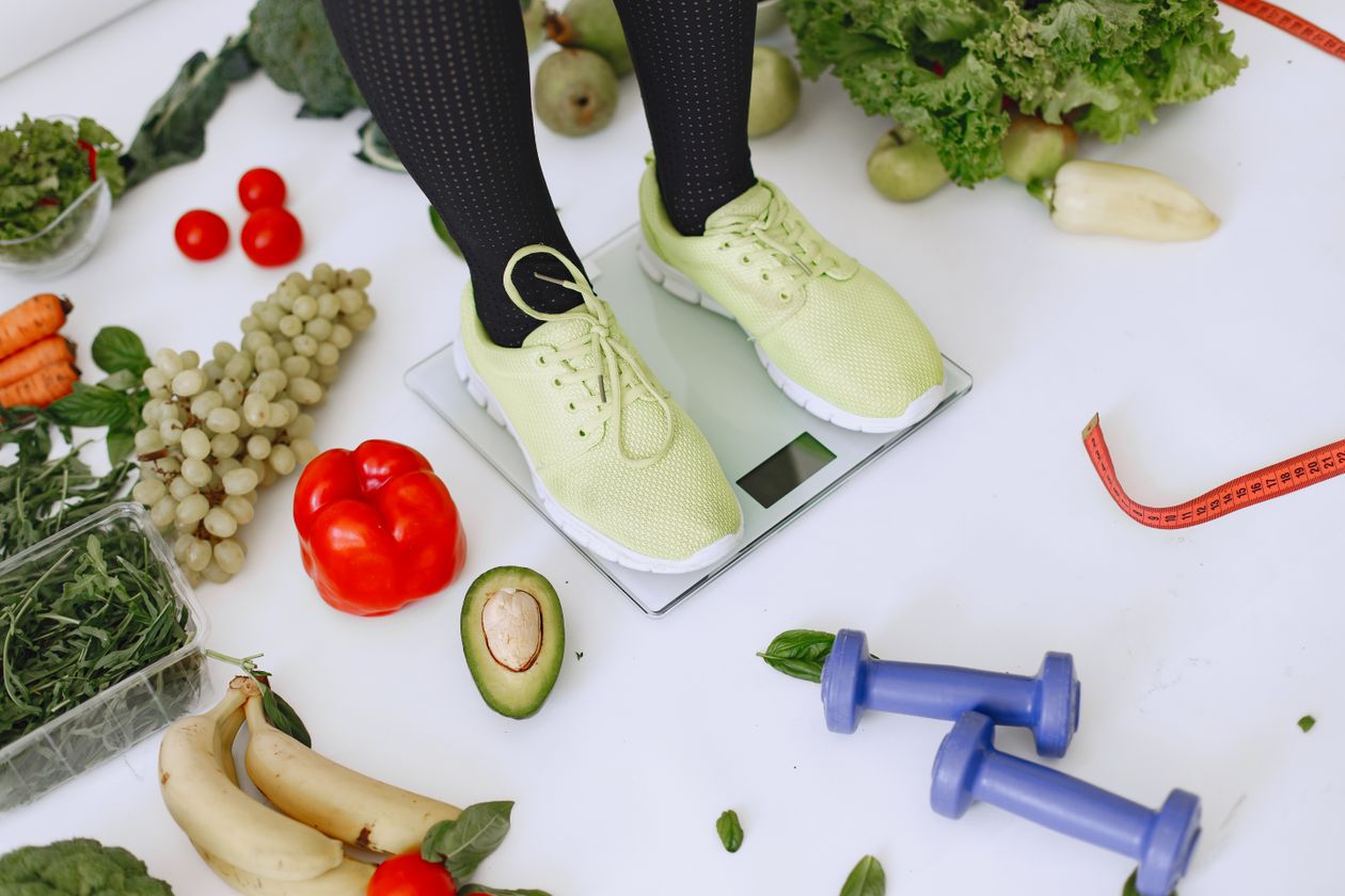 person standing on scales surrounded by fresh produce and fitness equipment symbolizing healthy weight management