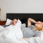 Man snoring on his back in bed while his partner covers her face, a common reason couples look for a butterfly pillow for snoring