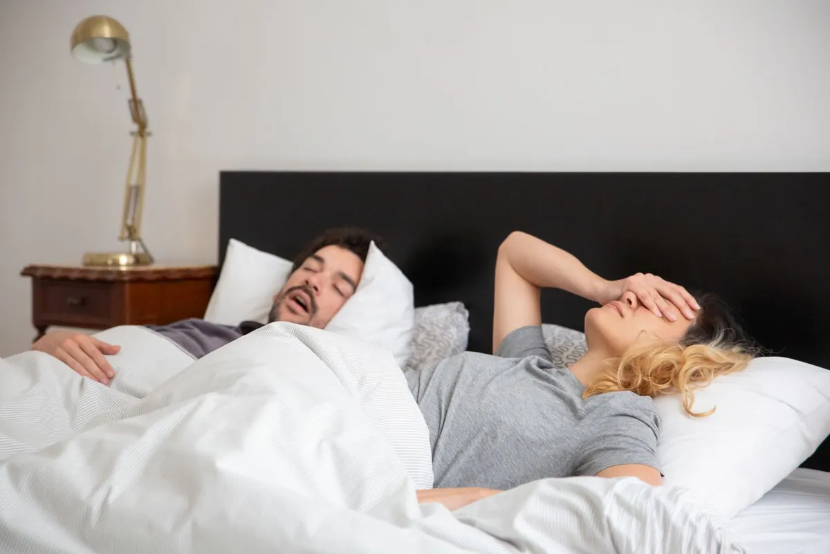 Man snoring on his back in bed while his partner covers her face, a common reason couples look for a butterfly pillow for snoring