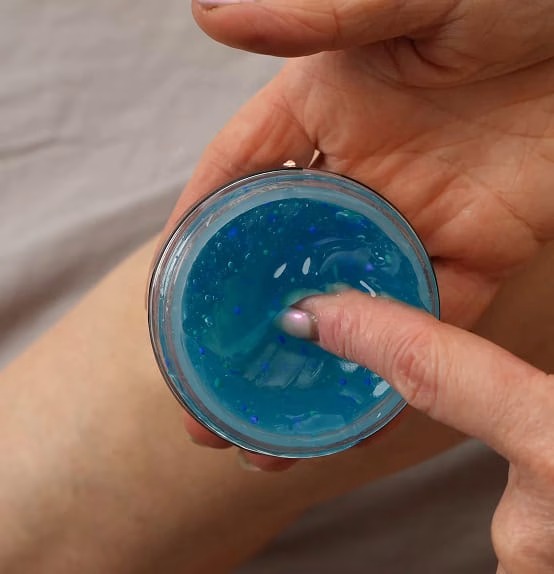 Hand holding an open jar of Kinzeno massage gel over a knee, taking gel on one finger to apply